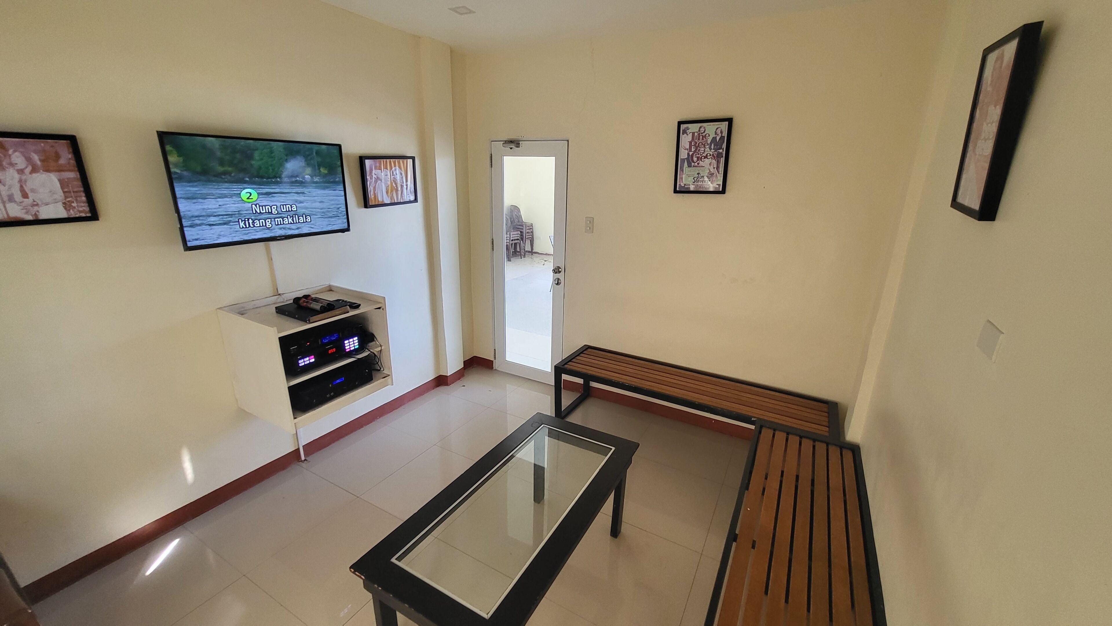 Una sala de estar con un televisor de pantalla plana montado en la pared, un banco de madera, una mesa de centro con superficie de vidrio y una puerta que da al exterior.