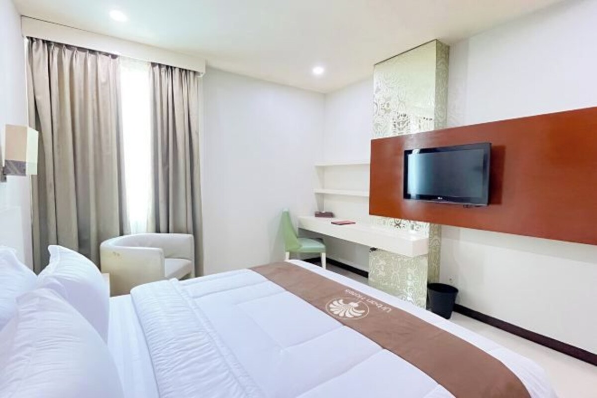A hotel room with a bed, a desk, a chair, a television mounted on the wall, and a window with curtains.