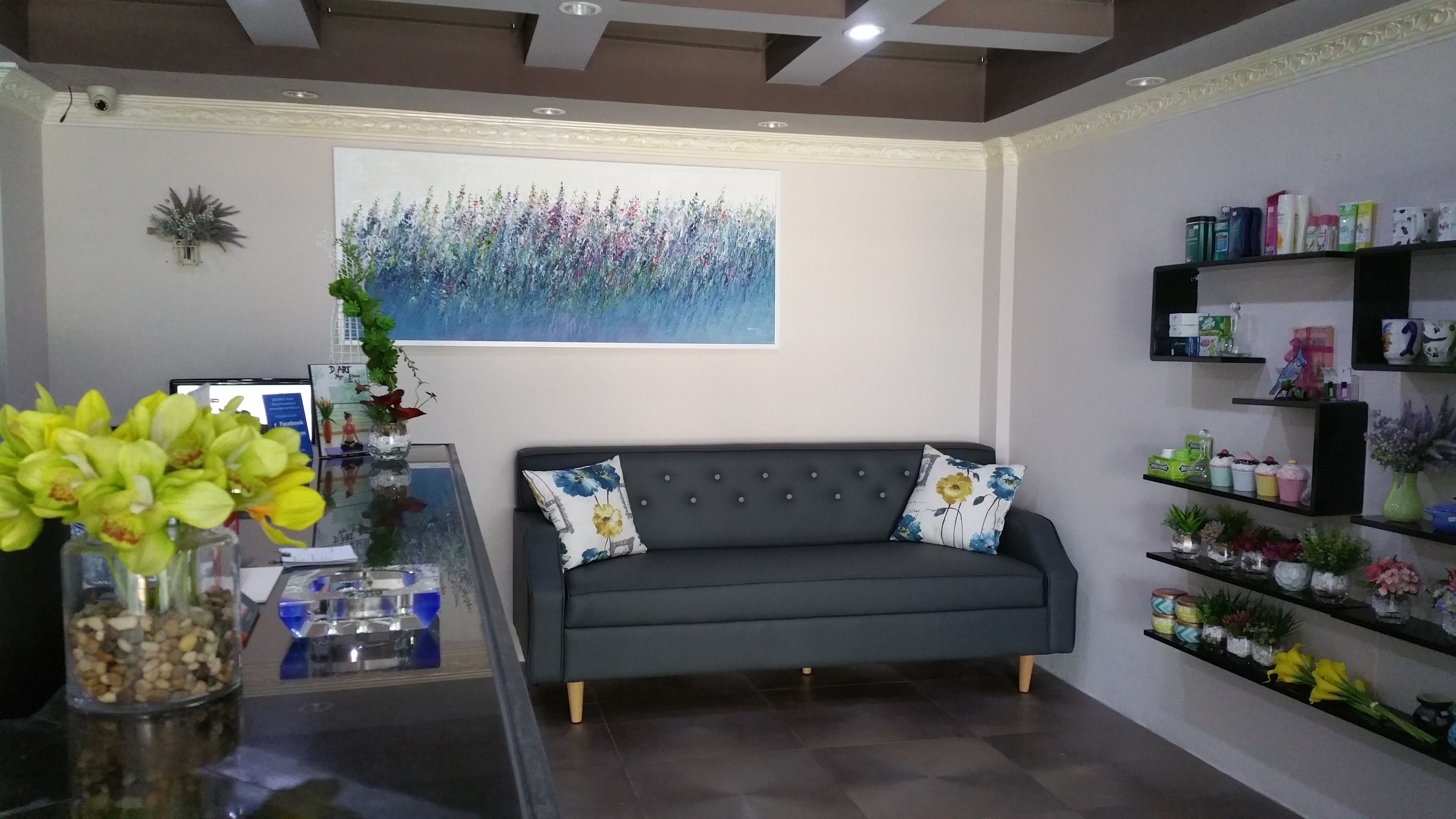 A modern living room with a grey sofa, decorative pillows, a glass coffee table, and a wall-mounted shelf with various items.