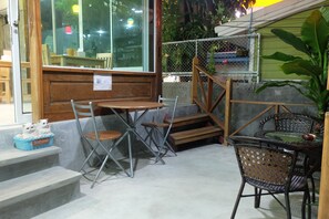 An outdoor seating area with metal chairs and tables, a wooden bench, and a wicker chair. There is a small basket with two cats and a welcome sign.
