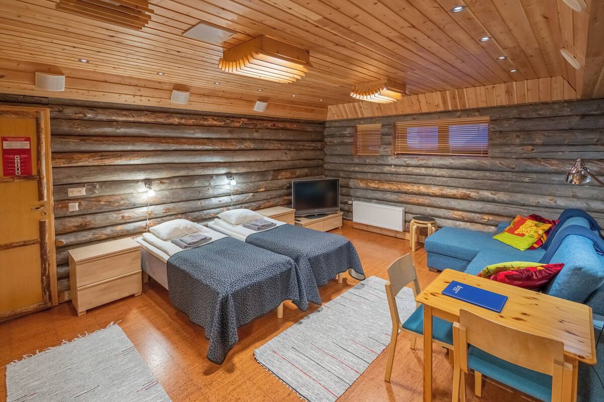 Interno di una baita di legno con due letti, una televisione, un divano e un tavolo in legno.