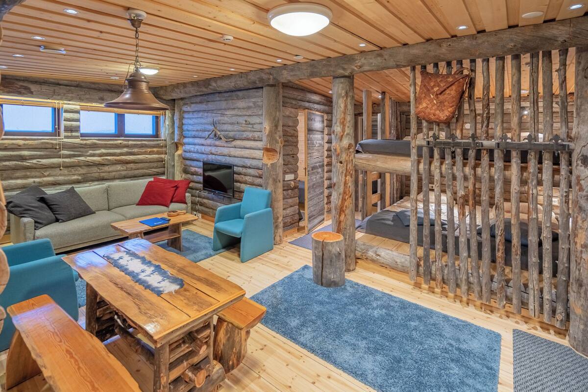 Interno di una baita con una poltrona, sedie blu, un tavolo in legno e una TV.