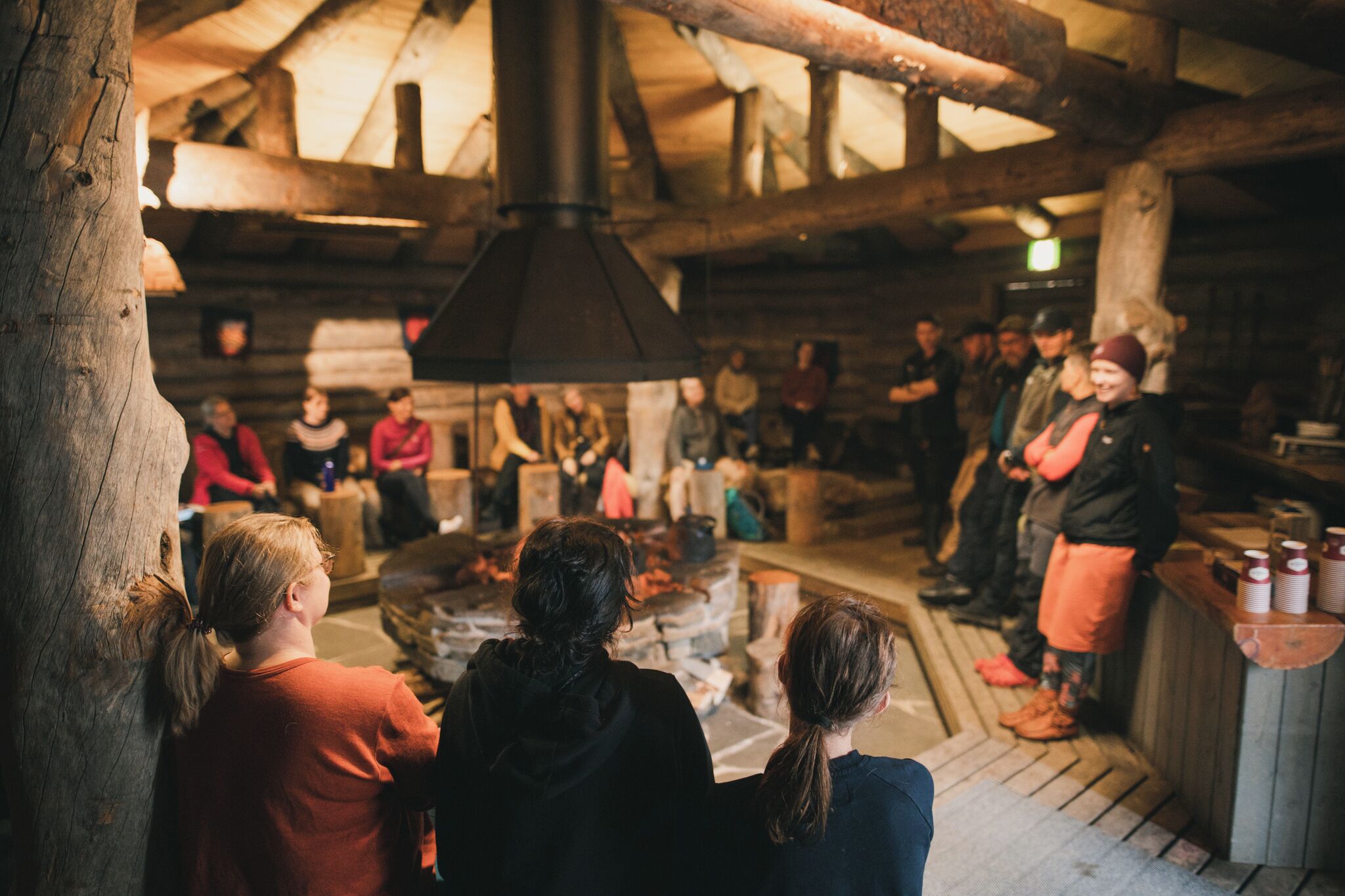 A rustic interior with a group of people gathered around a central fire, wooden beams on the ceiling, and a stone fireplace.