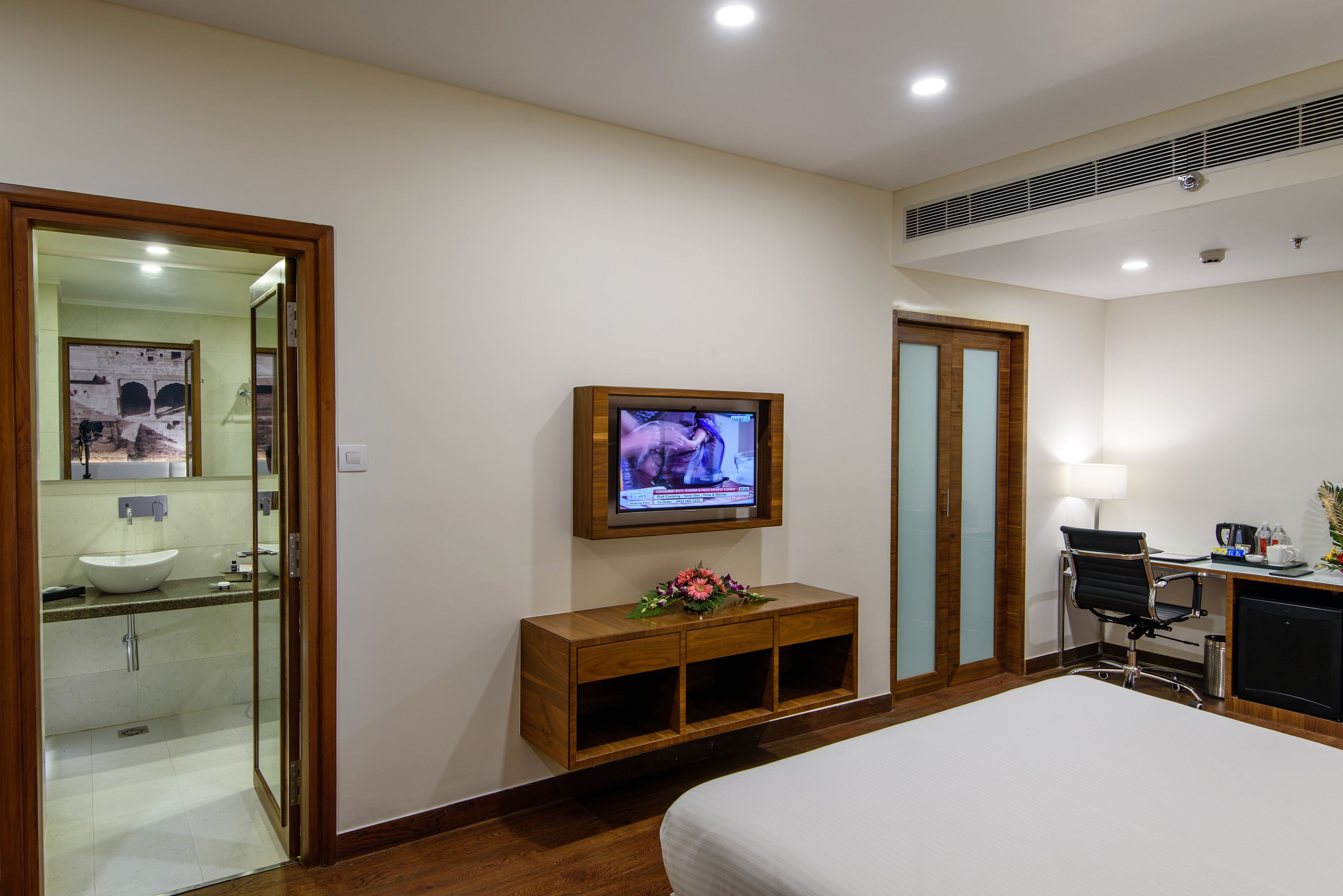 A hotel room with a bed, a TV mounted on the wall, a desk with a chair, and a wooden cabinet with a flower arrangement.