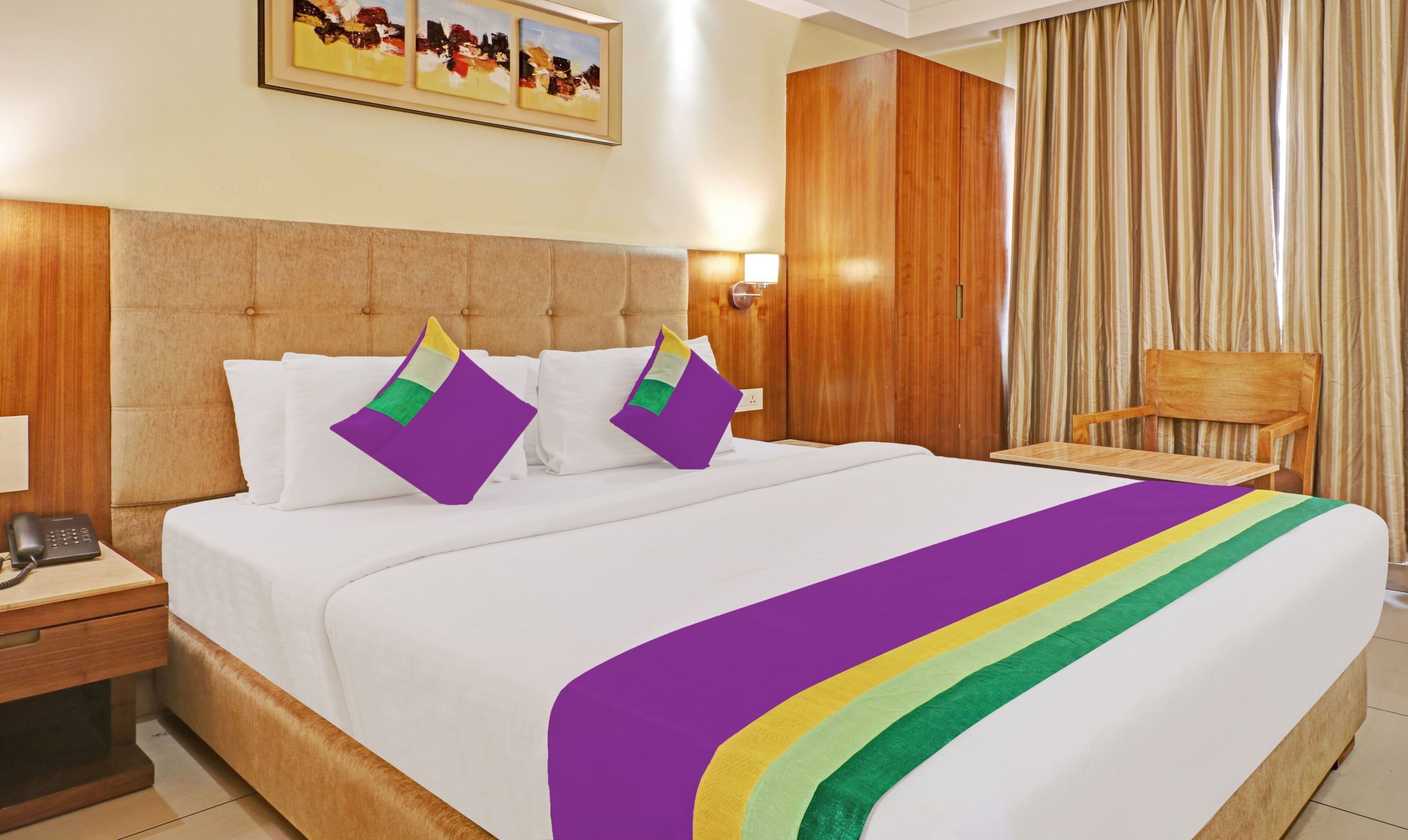 Een hotelkamer met een groot bed, twee kussens, een nachtkastje, een houten stoel, een houten garderobe en een schilderij aan de muur.
