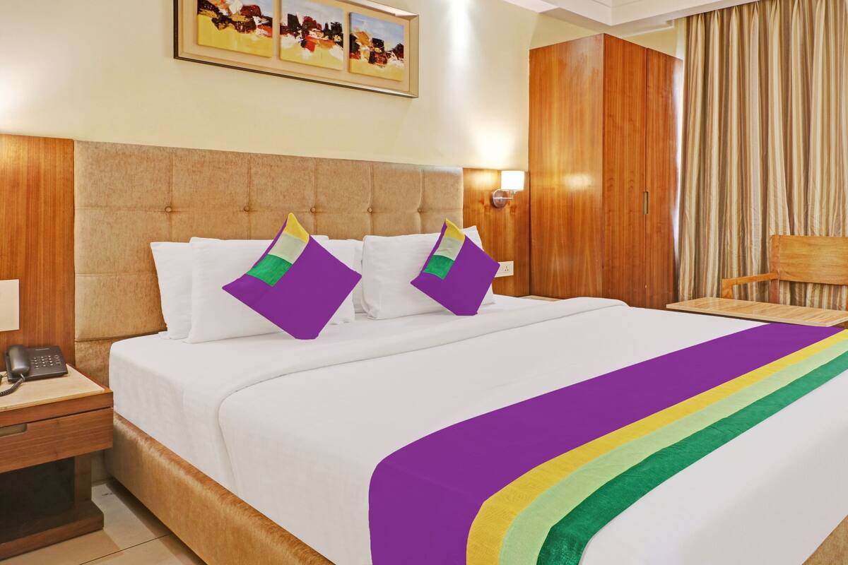 Quarto de hotel com uma cama grande, duas almofadas, uma mesa de cabeceira, uma cadeira de madeira, um guarda-roupa de madeira e um quadro na parede.