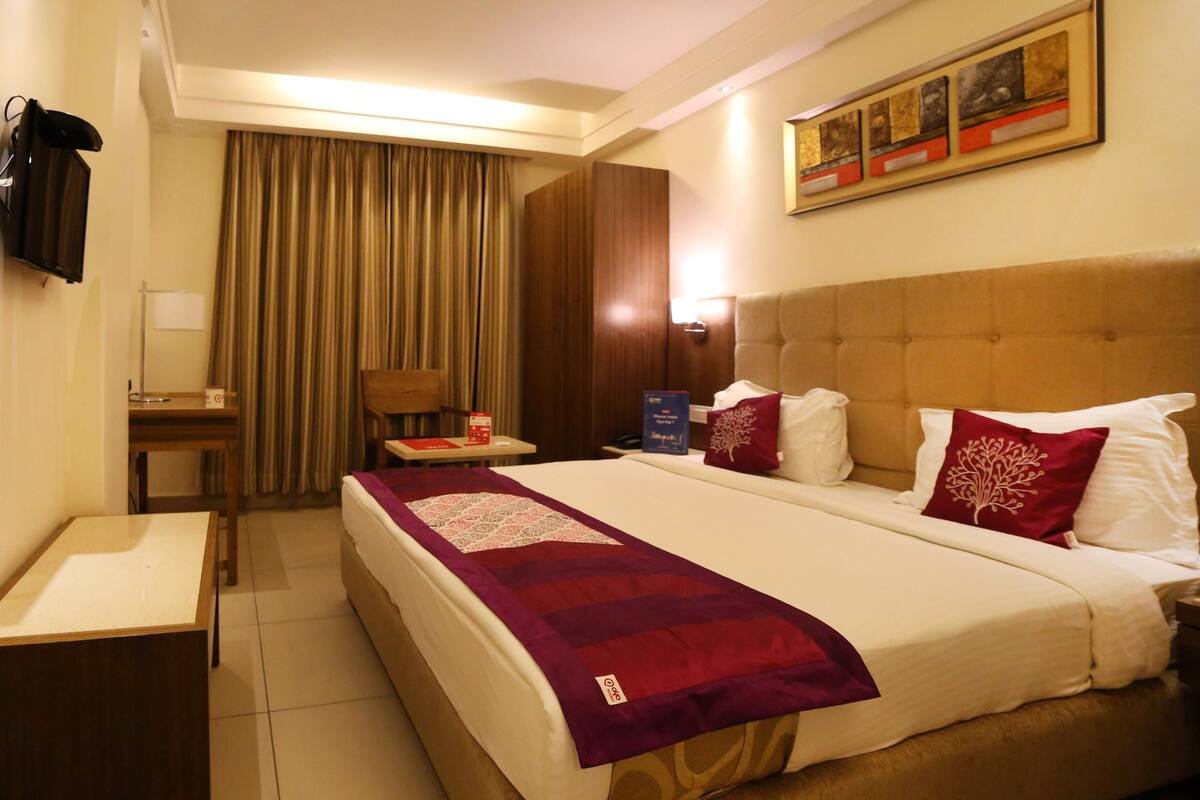 Quarto de hotel com uma cama grande, uma escrivaninha, uma cadeira, uma mesa de cabeceira e um quadro na parede.