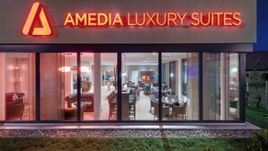 Un hôtel moderne avec une façade en verre, un espace de restauration visible avec des tables et des chaises, et une enseigne indiquant « AMEDIA LUXURY SUITES ».