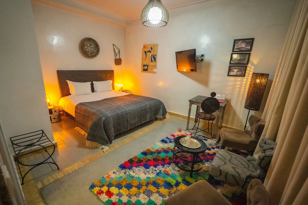 A hotel room with a bed, a small table, a chair, a colorful rug, and a wall-mounted TV.