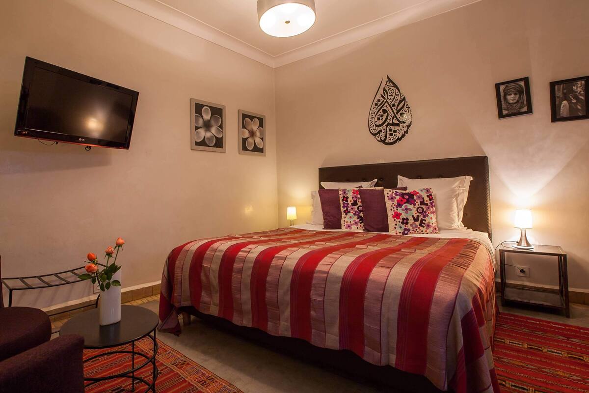 A hotel room with a bed, a TV mounted on the wall, a small table with a vase of flowers, and a chair.