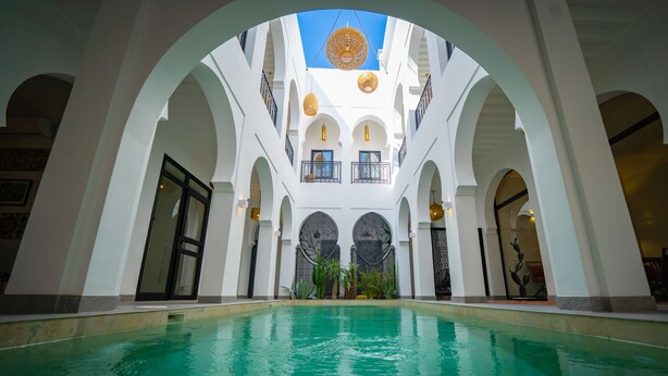 A pool area with a central water feature, surrounded by white arches and balconies with outdoor seating. - Riad Shanima & Spa (Marrakech)