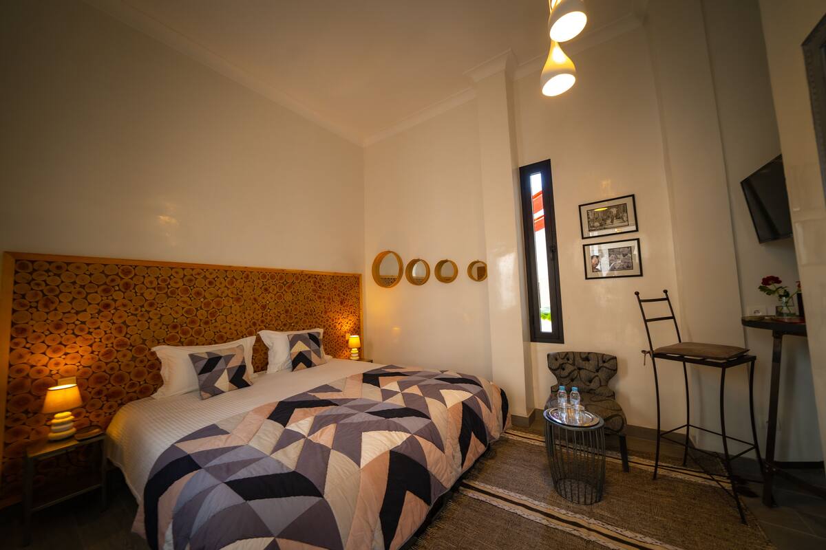 A hotel room with a large bed, a nightstand with a lamp, a chair, a small table with water bottles, and a TV on the wall.