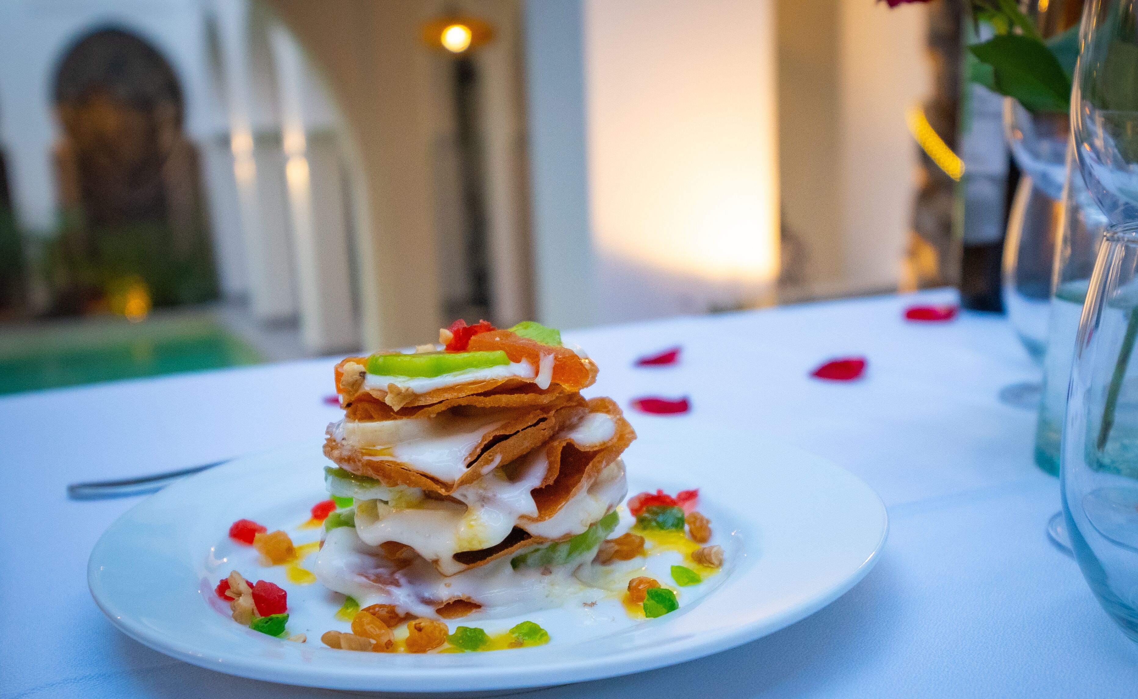 A dessert on a white plate with colorful toppings, set on a table with a glass and a vase in the background.