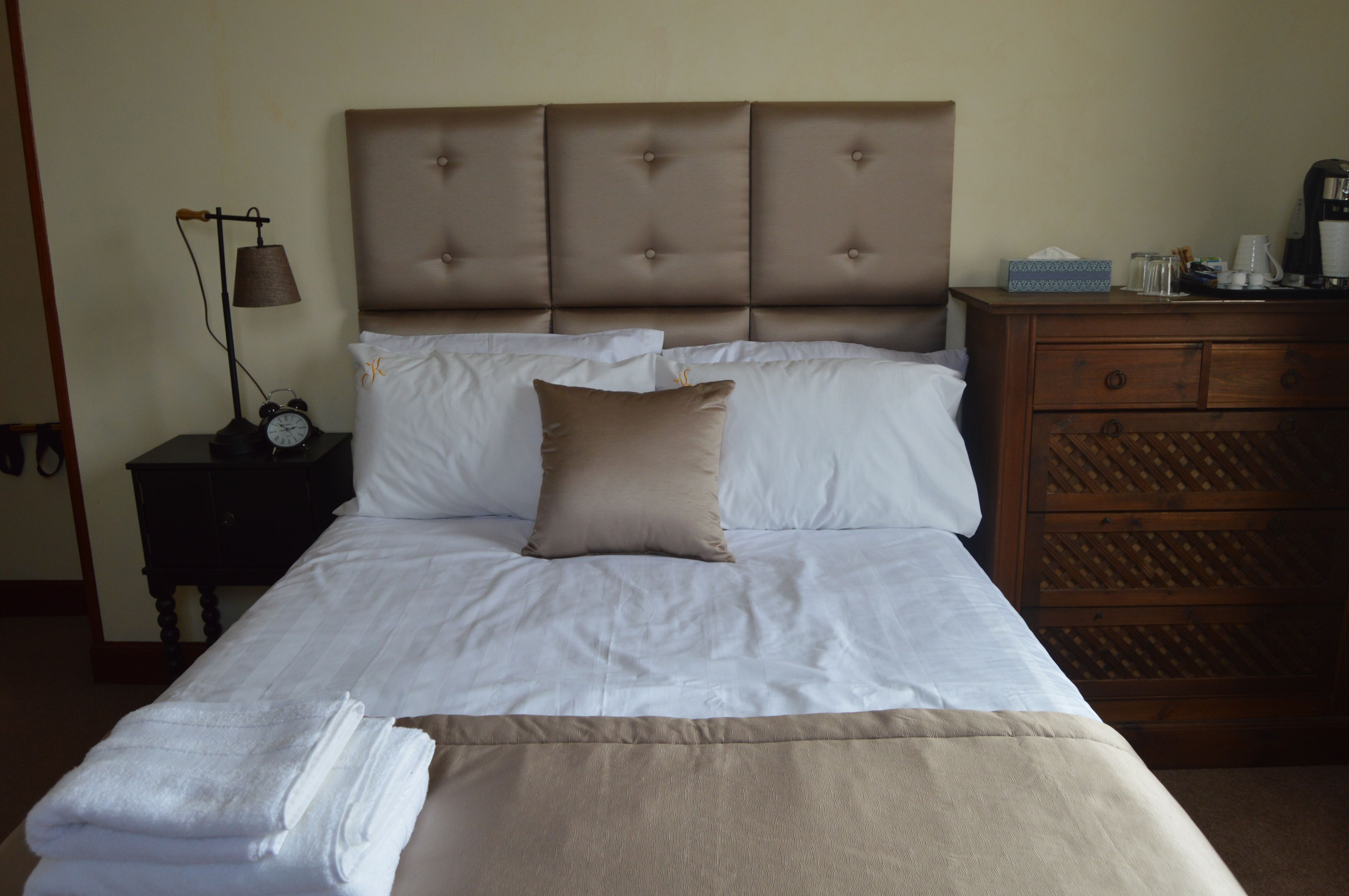 A neatly made bed with white linens and a brown headboard, a bedside table with a lamp and alarm clock, and a wooden dresser with a mirror.