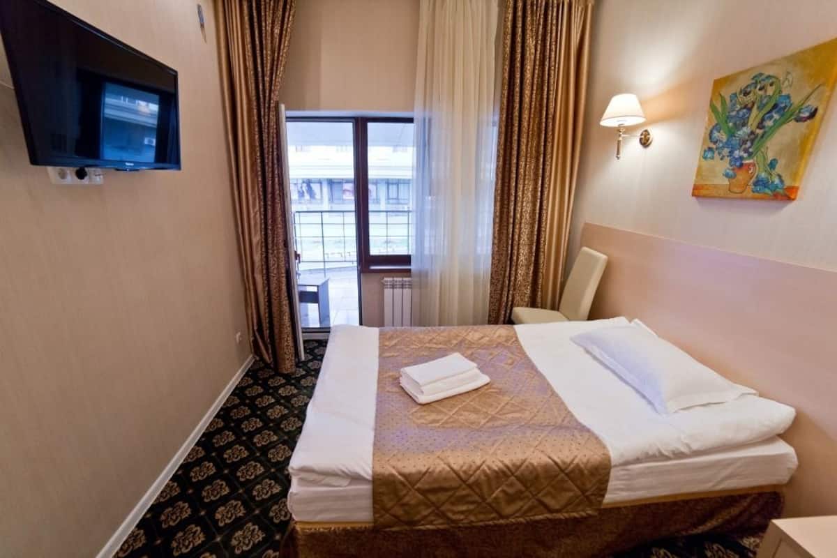 A hotel room with a bed, a TV mounted on the wall, a painting, and a window with curtains.