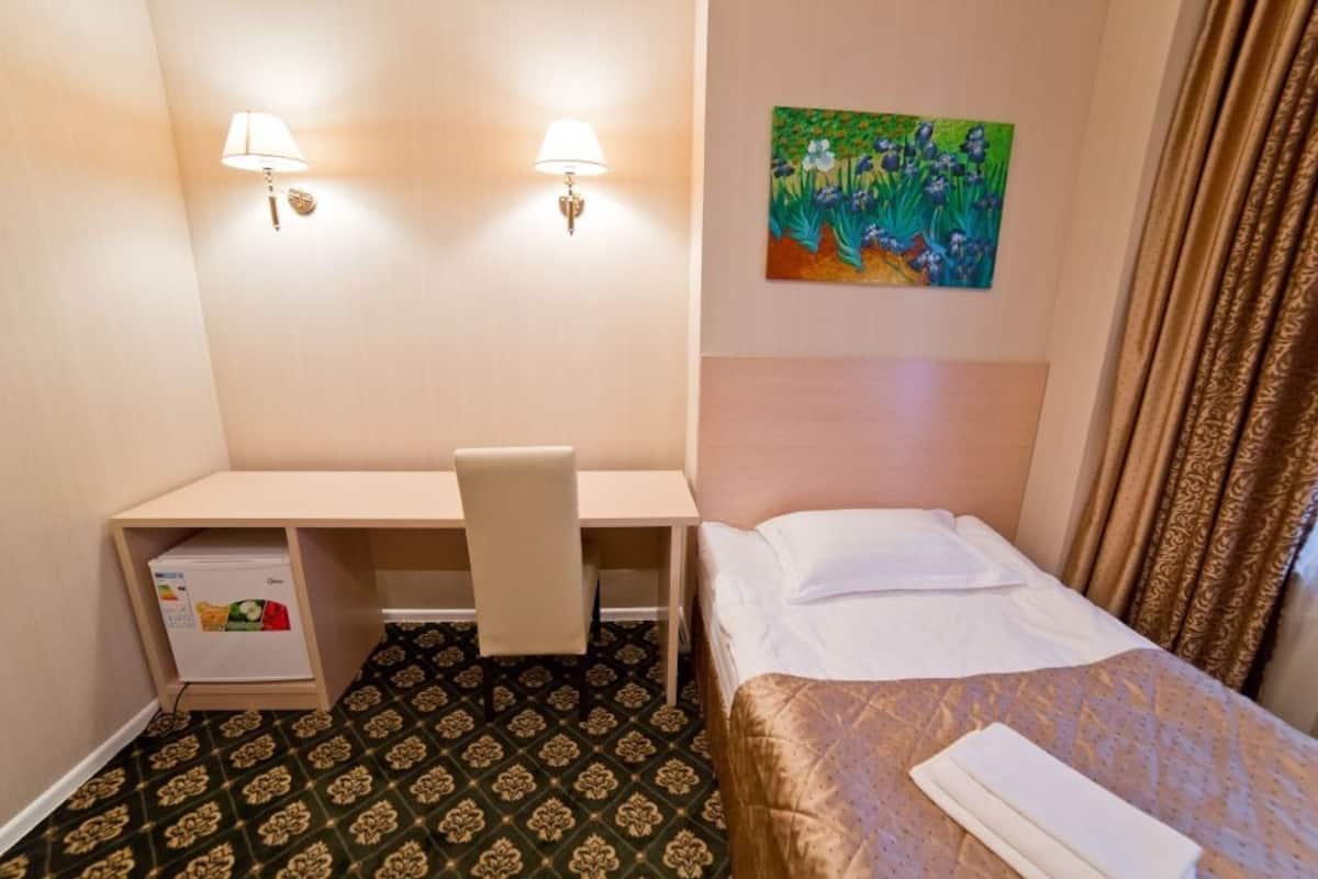 A hotel room with a bed, a desk, a chair, a small refrigerator, and a painting on the wall.