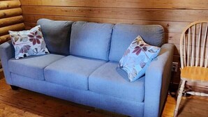 Interior de una cabaña de troncos con un sofá azul y dos almohadones, paredes de madera y una silla de madera.