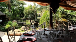 An outdoor dining area with a table set for a meal, surrounded by lush greenery and a thatched-roof structure.