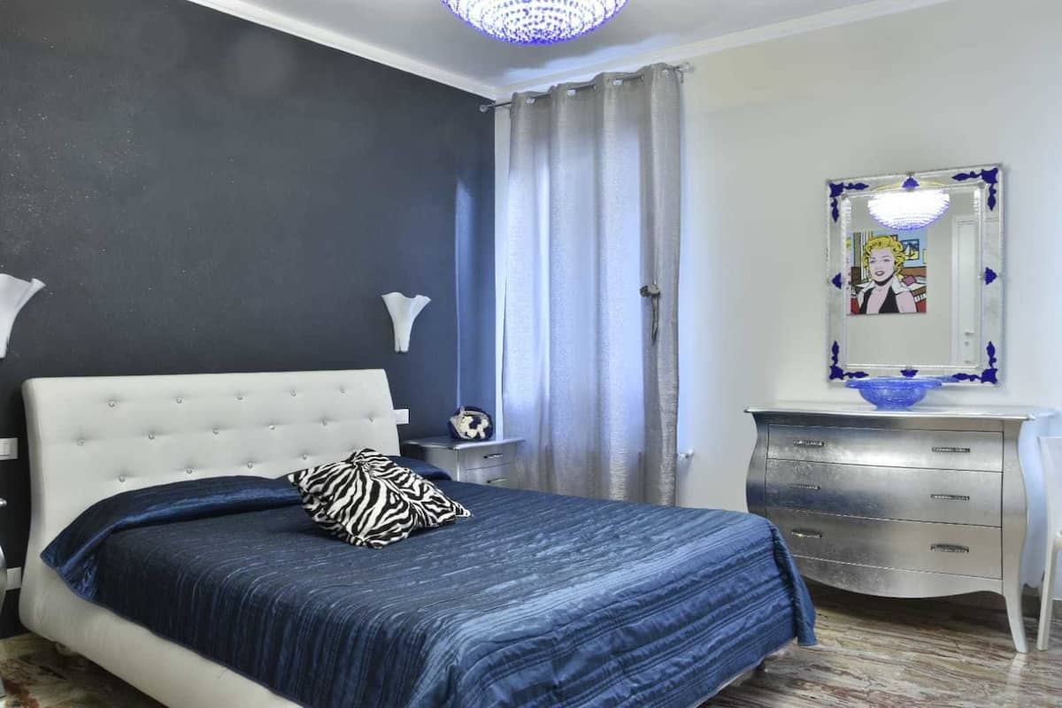 A bedroom with a tufted headboard bed, a blue bedspread, a zebra-patterned pillow, a white bedside table with a lamp, a dresser with drawers, and a mirror with a decorative frame.
