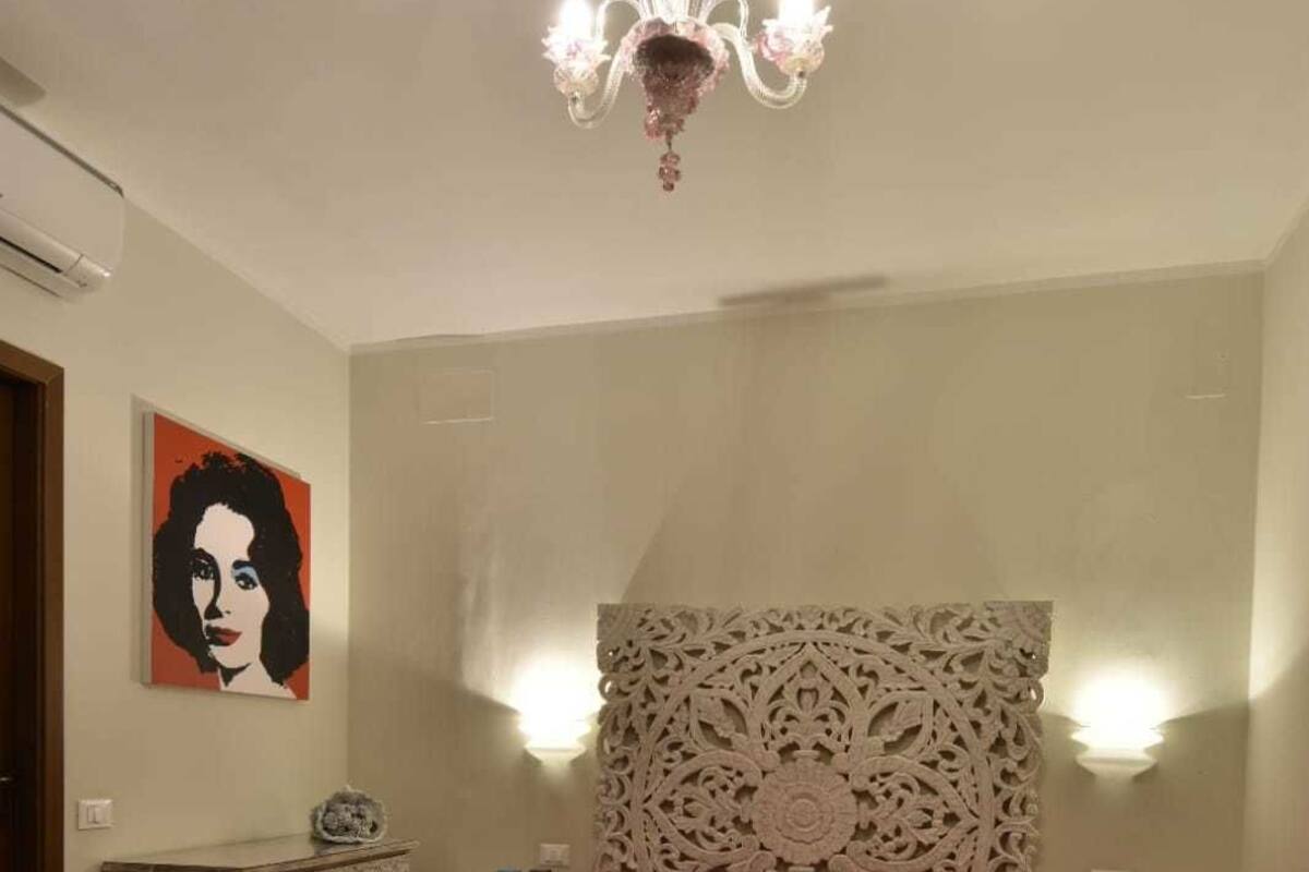 A bedroom with a large bed, a chandelier, a decorative headboard, a painting on the wall, and a dresser.