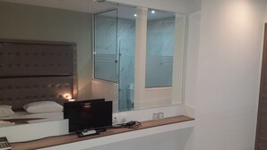 A modern bedroom with a large bed, a TV, a bedside table with glasses, and a mirror reflecting a bathroom. - Hotel Rex (Thessaloniki)