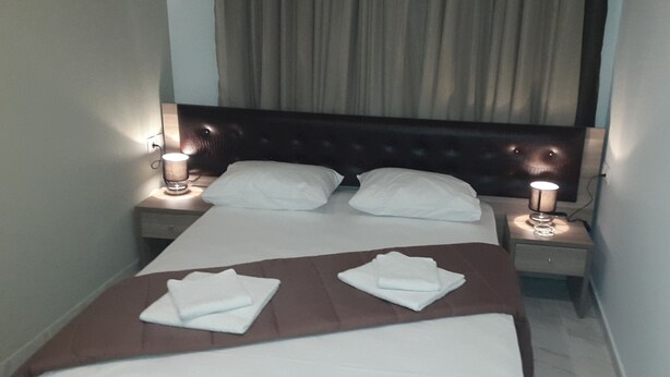 A neatly made bed with white linens and brown headboard, two bedside lamps, and a brown bedside table with a lamp. - Hotel Rex (Thessaloniki)