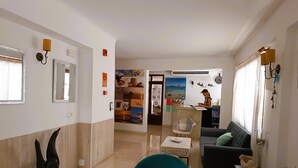 Una sala de estar moderna con un sillón verde azulado, un sofá negro y una mesita con adornos. Una mujer trabaja en un escritorio al fondo.