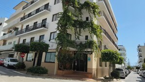 Un edificio de varios pisos con balcones, una entrada visible y coches estacionados a lo largo de la calle.