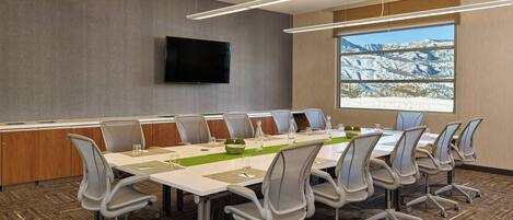 A conference room with a long table, chairs, a flat-screen TV, and a large window with a mountain view.
