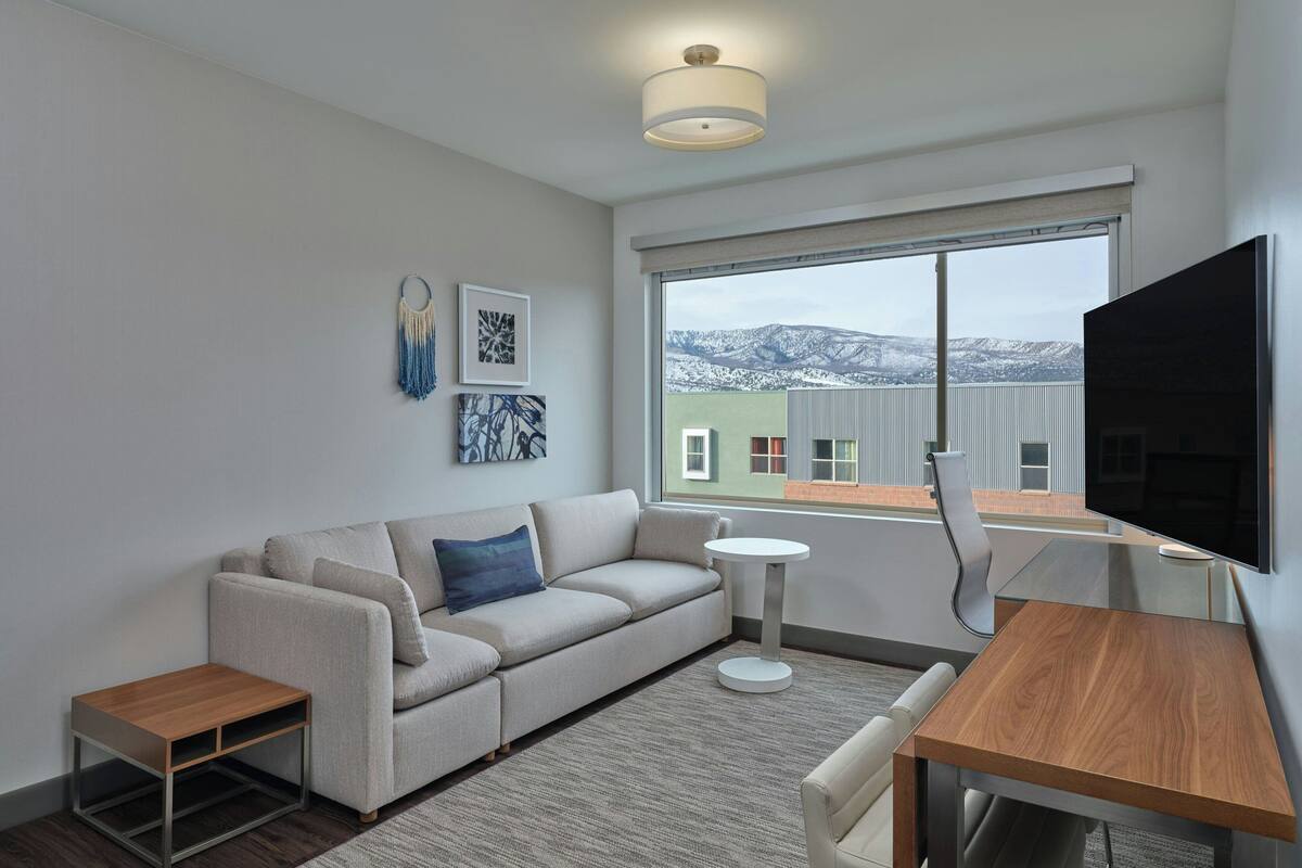 A modern living room with a sofa, a small table, a TV, and a large window with a view of buildings and mountains.