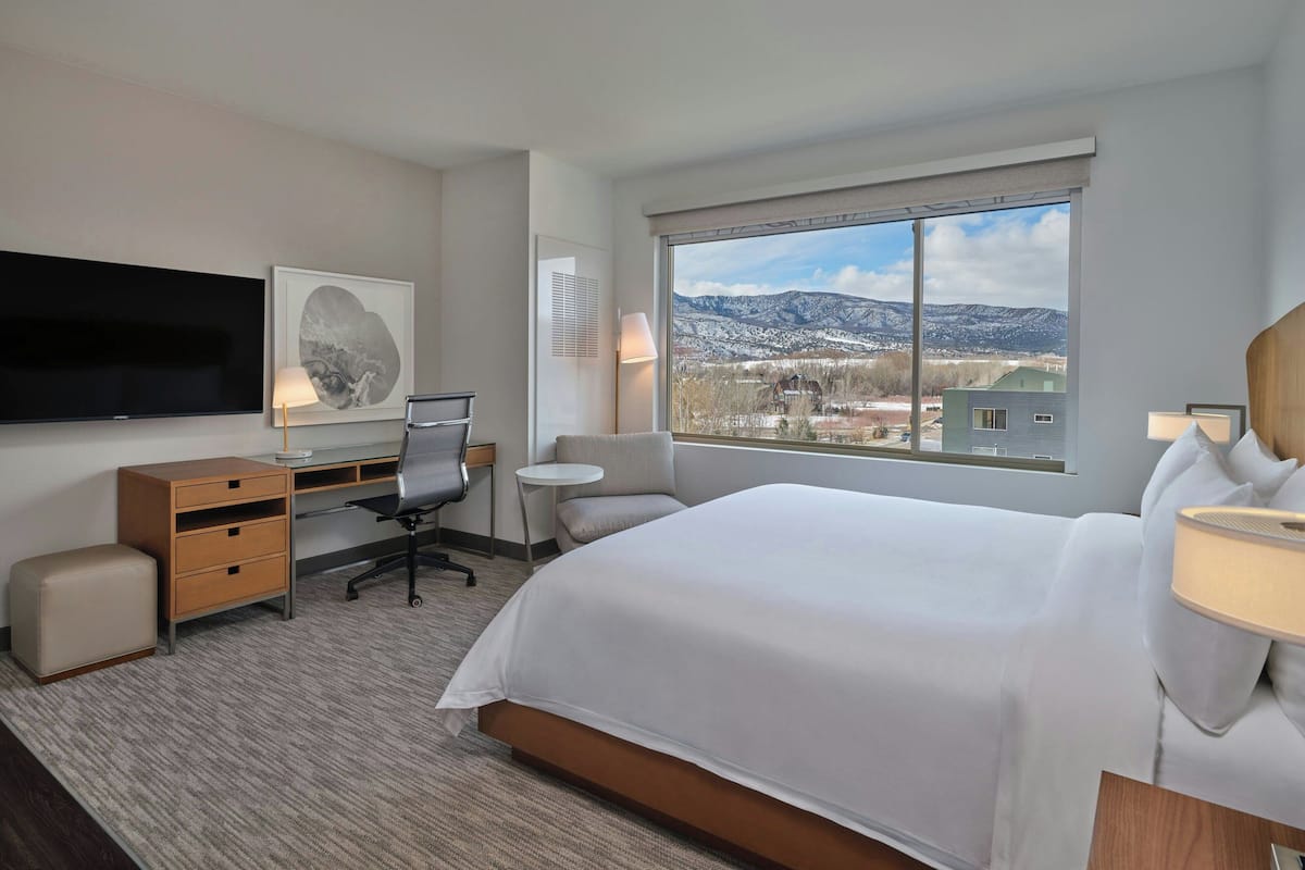A hotel room with a large bed, a desk with a chair, a television, a window with a mountain view, and a small table with a lamp.