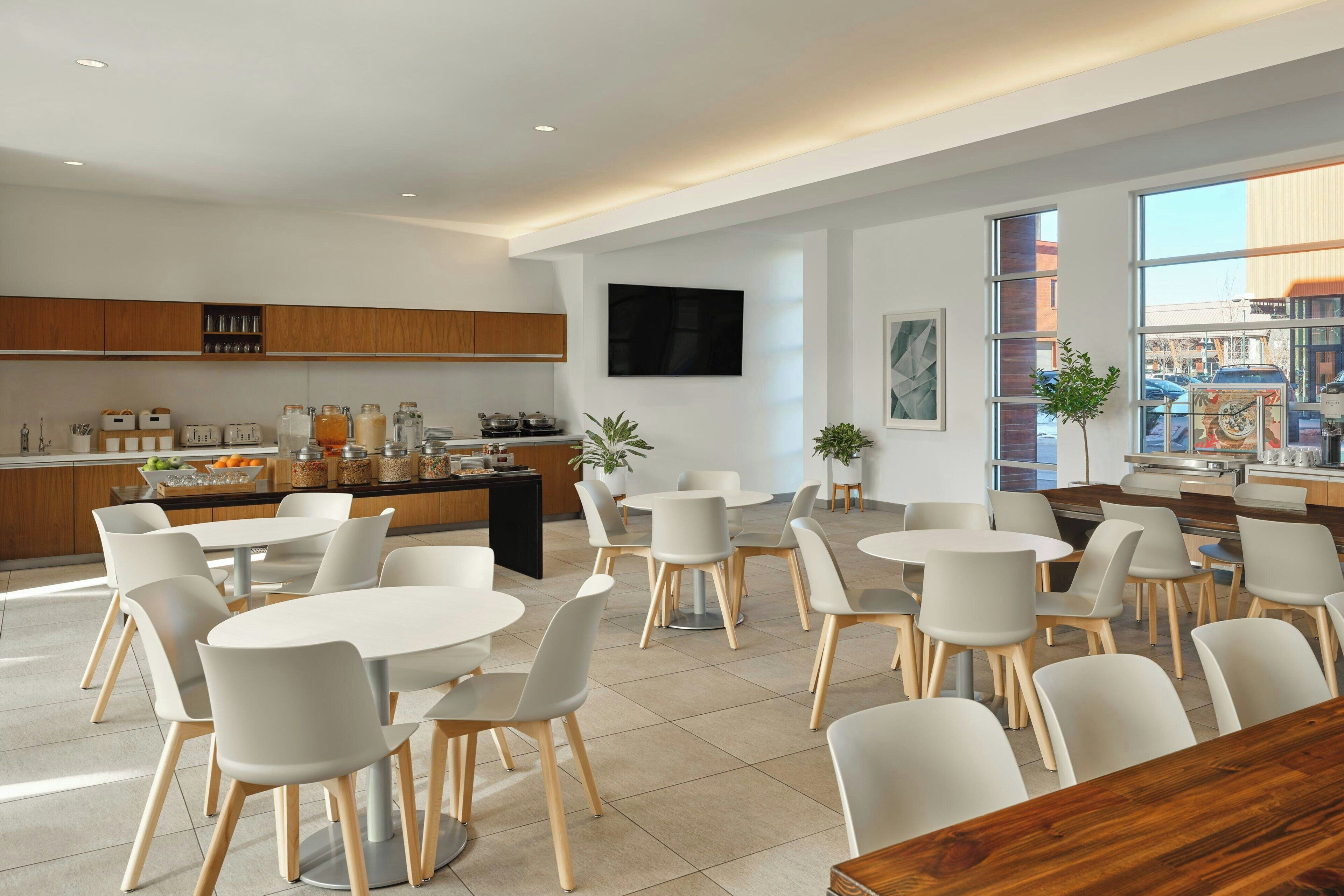 Un comedor moderno con mesas y sillas blancas, una cocina al fondo y ventanas amplias con vistas al exterior.