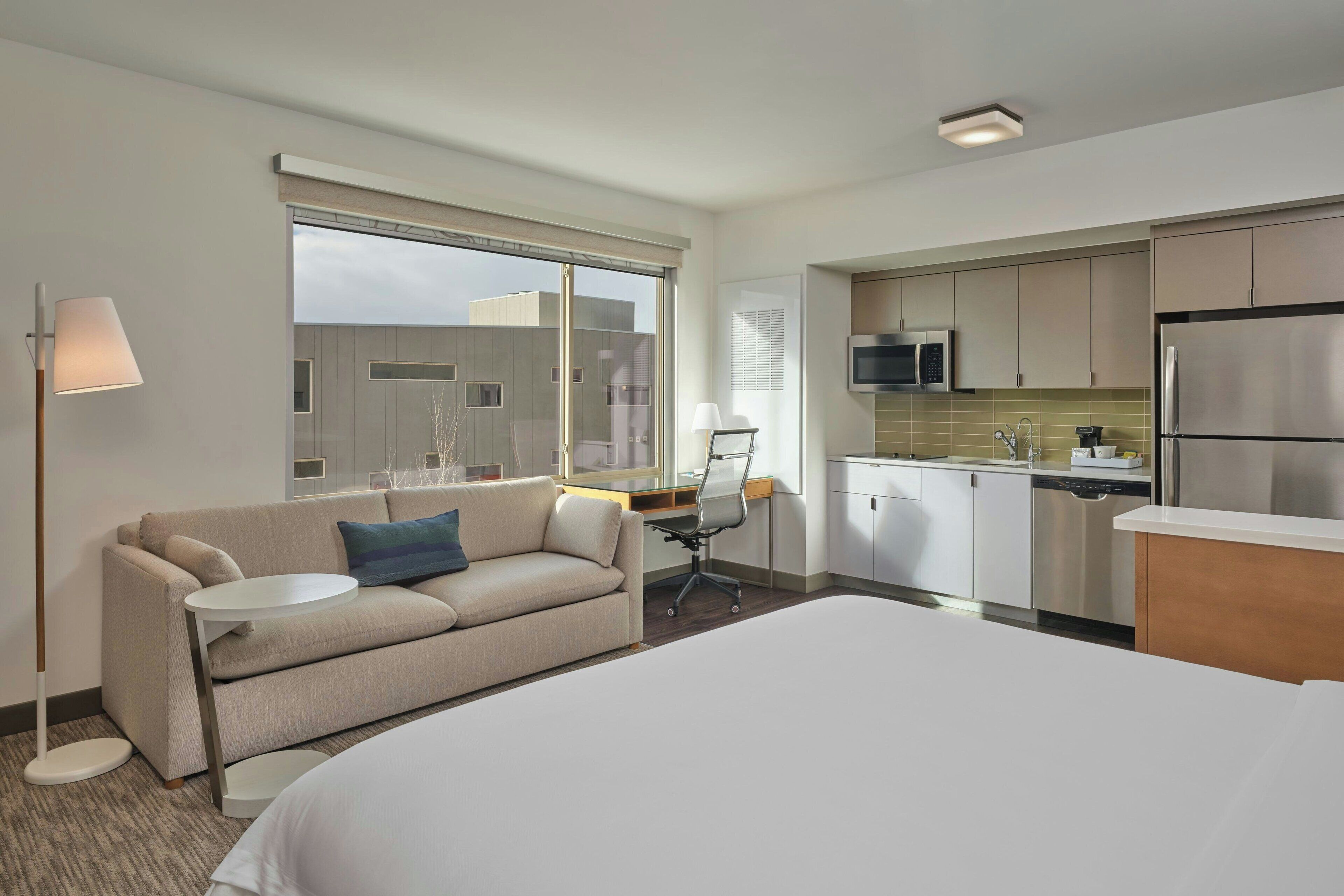 A hotel room with a bed, a sofa, a desk, a lamp, a TV, and a kitchenette.