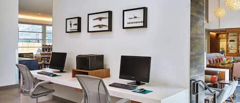 A modern office space with a long white desk, two computers, and two office chairs. There are framed pictures on the wall and a dining area in the background.