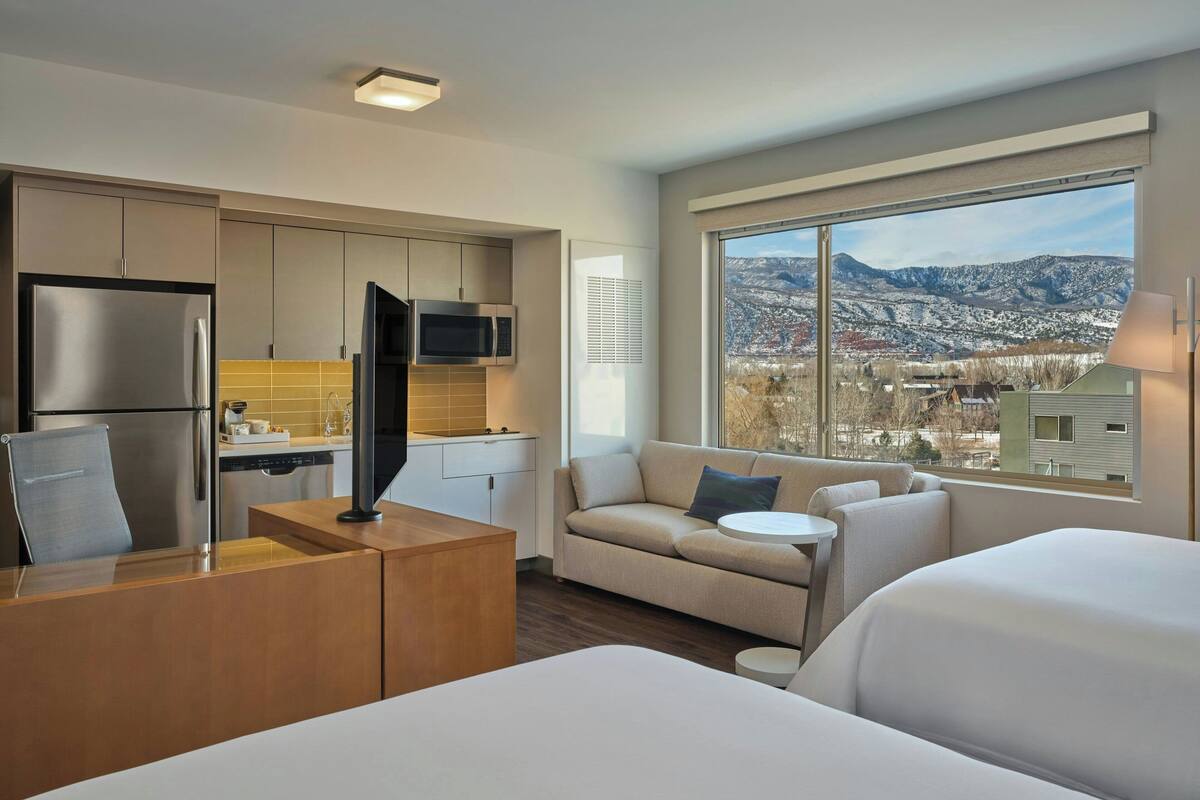 A modern hotel room with a kitchenette, a sofa, a bed, and a large window offering a city and mountain view.