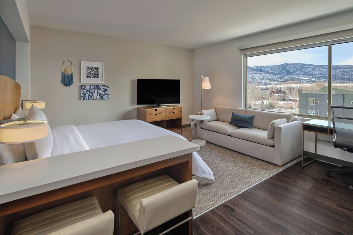 A hotel room with a bed, a sofa, a desk, a TV, and a large window offering a mountain view.