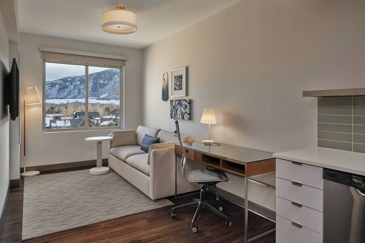A modern living room with a sofa, desk, and dining area. A large window offers a view of a snowy landscape.
