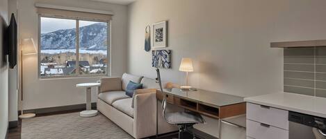 A modern living room with a sofa, desk, and dining area. A large window offers a view of a snowy landscape.