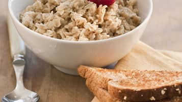A bowl of oatmeal with strawberries on a wooden table, accompanied by a spoon and slices of bread on a napkin.