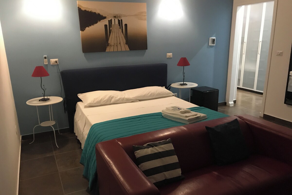 A hotel room with a bed, a red leather sofa, a painting on the wall, and a red lamp on a side table.