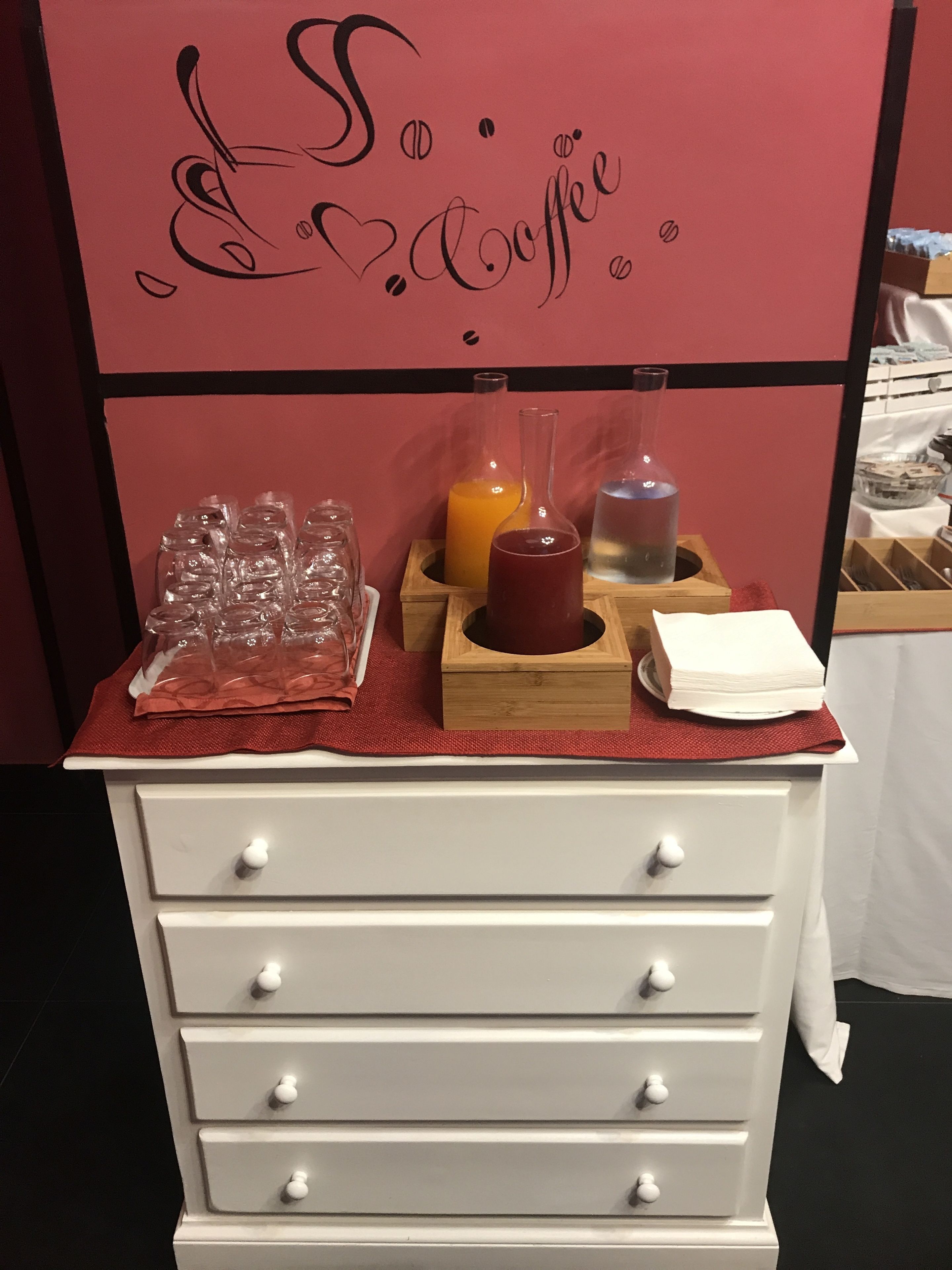 Eine weiße Kommode mit Schubladen, eine rote Unterlage sowie verschiedene Gegenstände, darunter ein Glasbehälter, eine Orangensaftflasche, ein Glas Wasser und ein Servietttuch.
