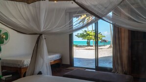 A room with a bed, a bedside table, a fan, and a view of a beach with palm trees and the ocean. - Cristal Resort (Paje)