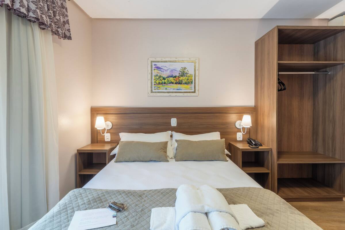 A hotel room with a large bed, two bedside tables with lamps, a wooden wardrobe, and a framed landscape painting on the wall.