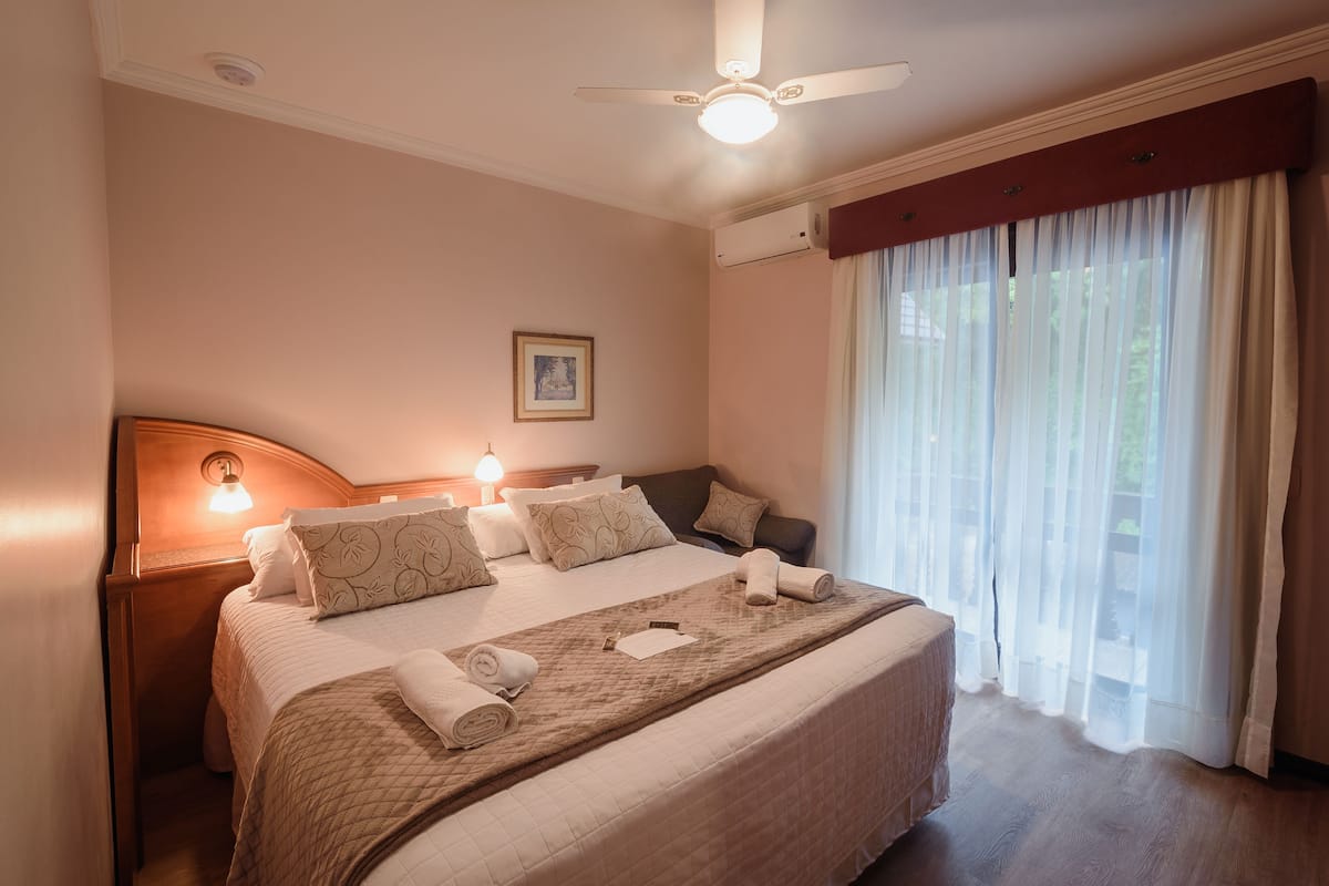 A neatly made bed with a quilt, pillows, and a blanket rolled at the foot. There is a ceiling fan, a wall-mounted light, and a window with sheer curtains.