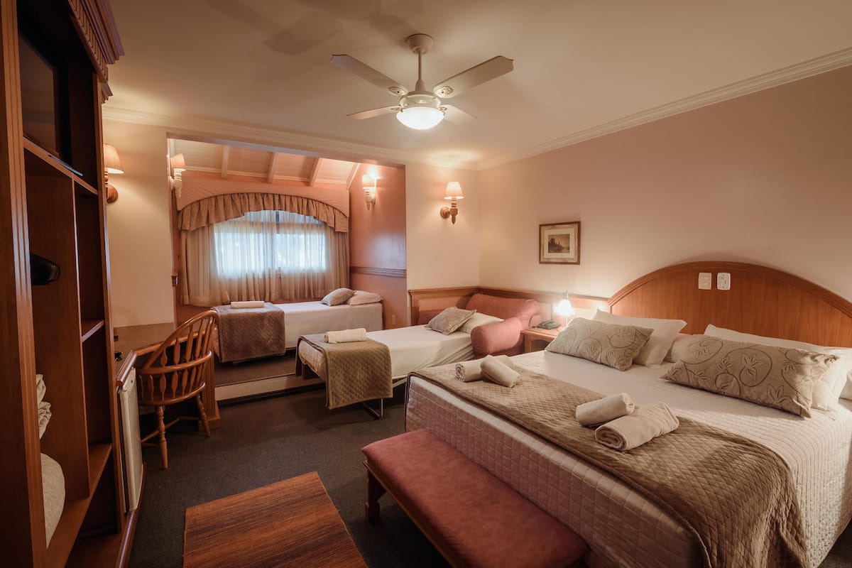 A hotel room with two single beds, a double bed, a wooden wardrobe, a ceiling fan, and a window with curtains.