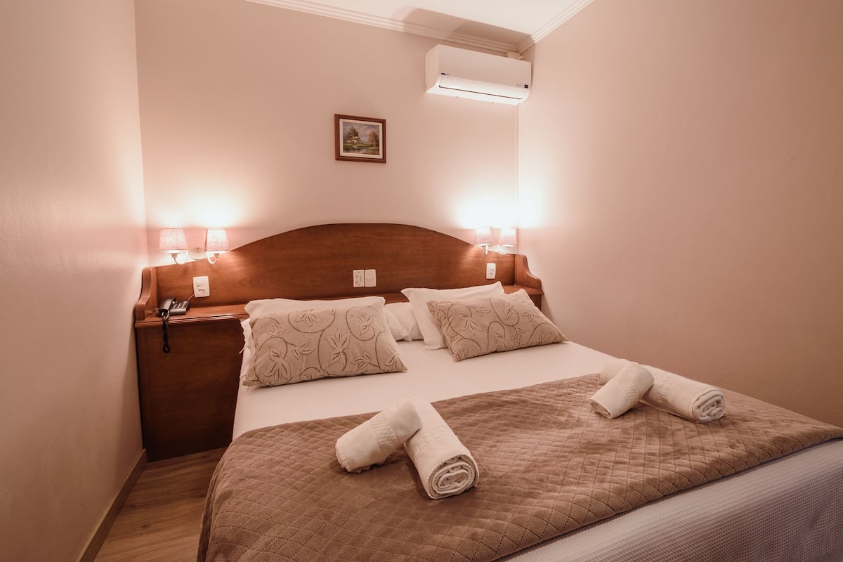 A neatly made bed with a quilted brown cover, two pillows with a floral pattern, and two rolled towels. There is a bedside table with a lamp and a phone.