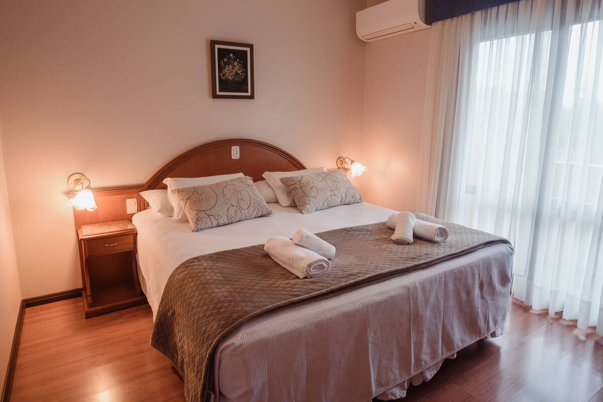 A neatly made bed with a brown quilt, two pillows, and two rolled towels. There is a nightstand with a lamp and a wall-mounted picture.