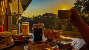 Una mesa junto a una ventana con vista al atardecer, con un vaso de jugo, una taza y bollería.