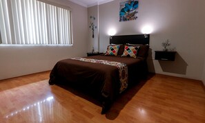 Un dormitorio con una cama grande, piso de madera, una ventana con persianas, una mesita de noche, un estante en la pared y un cuadro.