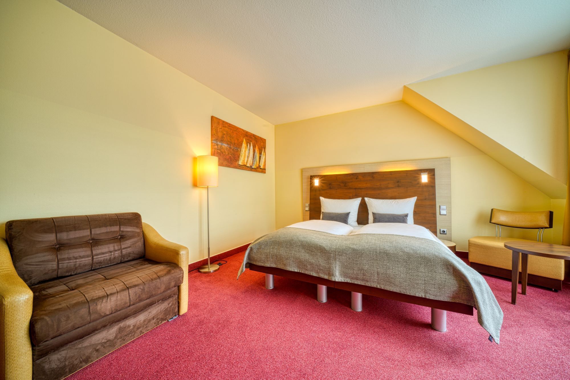 A hotel room with a bed, a sofa, a floor lamp, and a painting on the wall.
